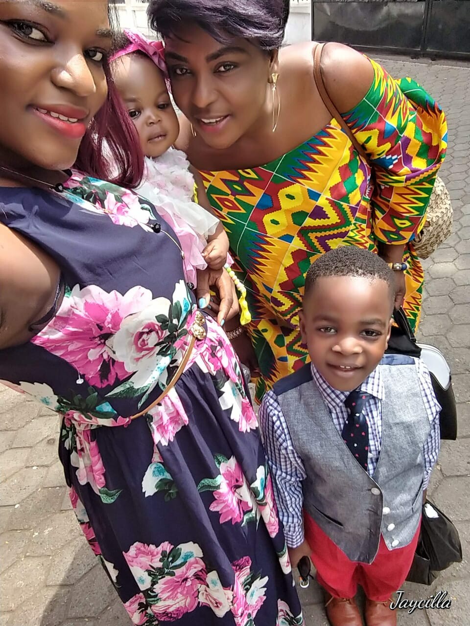 Three people posing for a selfie: a pregnant woman in a purple floral dress, a woman in a bright geometric-pattern dress, and a smiling boy in a gray vest and tie on a sidewalk.