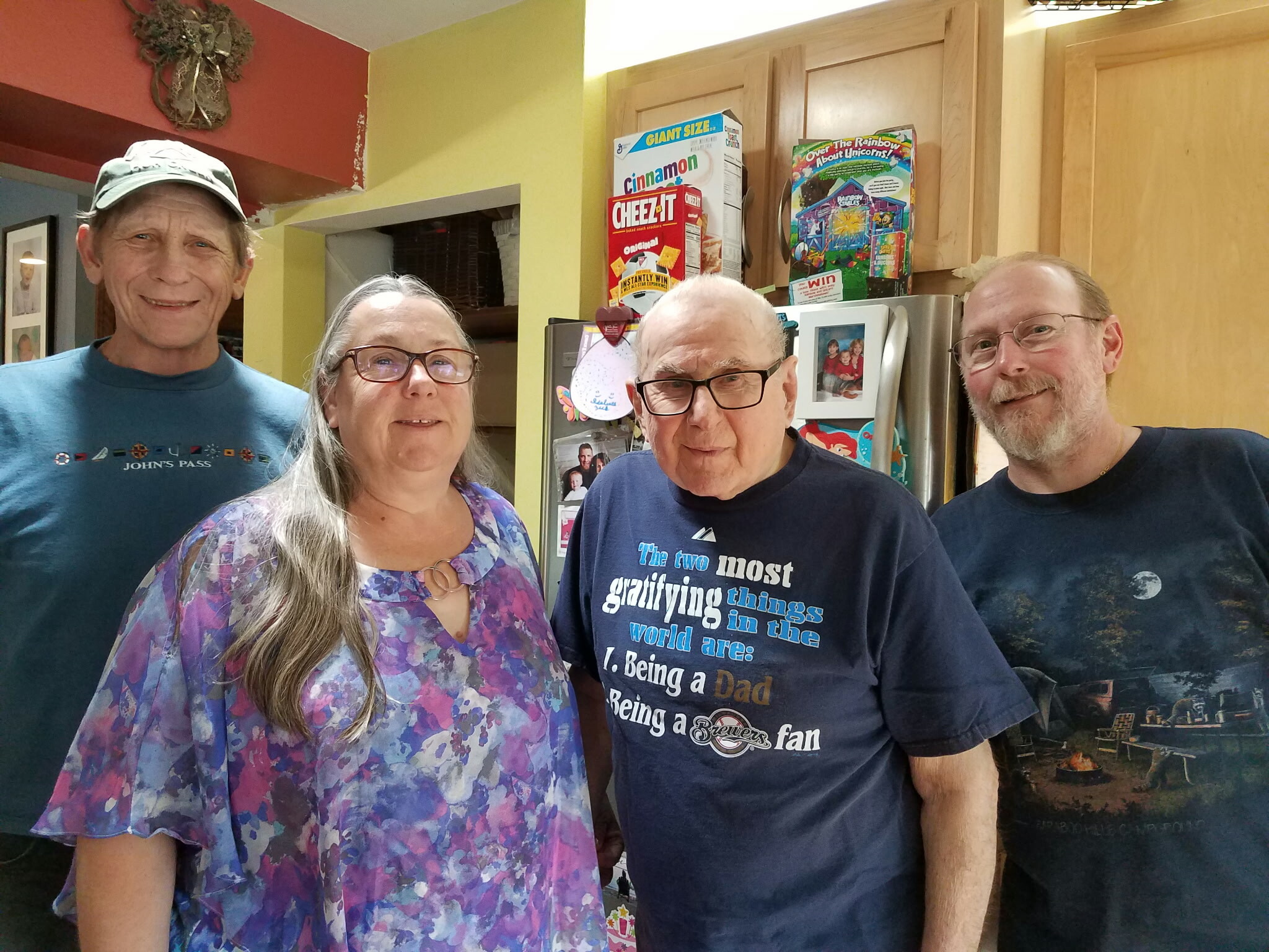 Four adults smile for a casual group photo in a home kitchen, with cereal boxes on the counter behind them and colorful walls on the left.