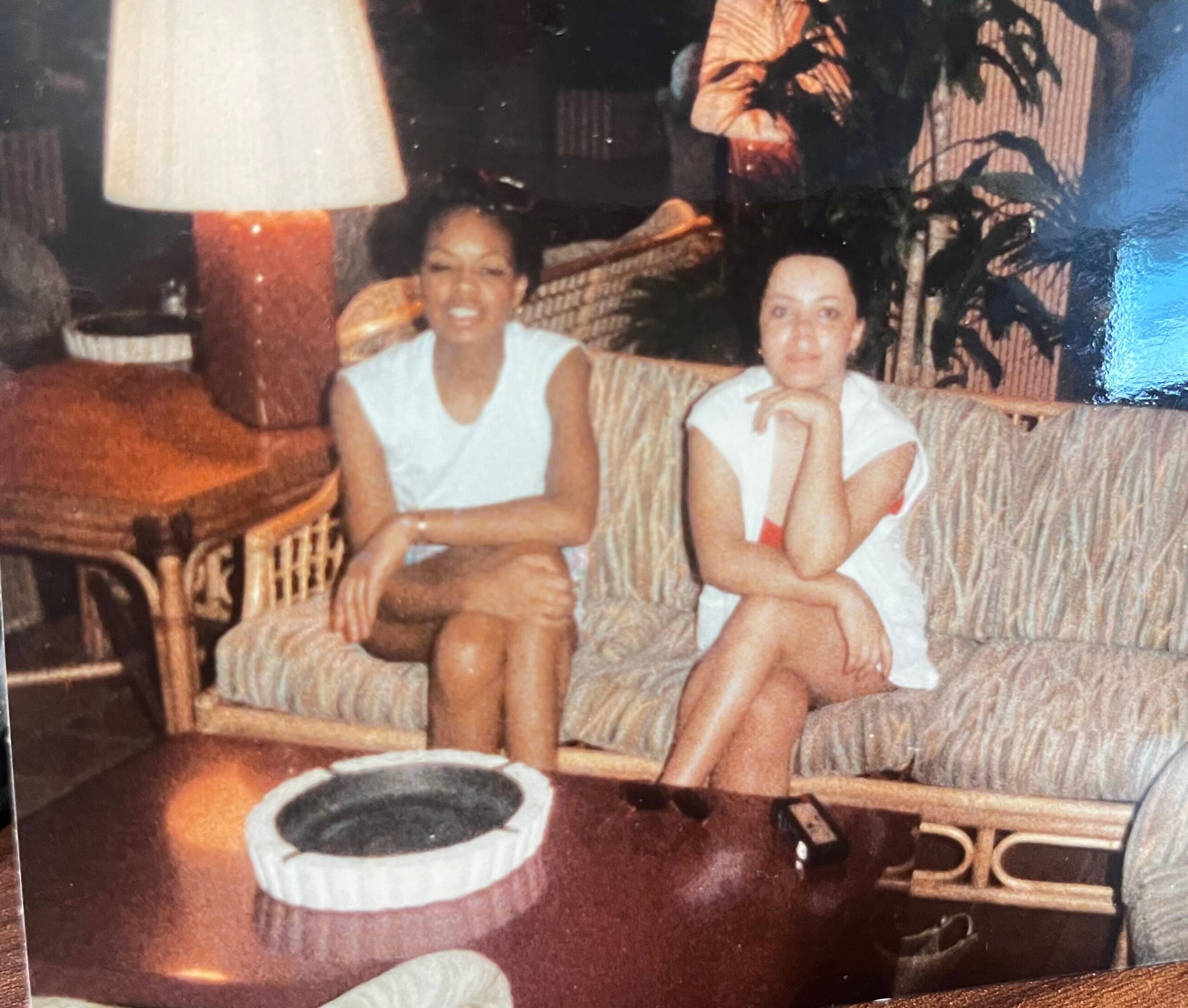 Two women sit on a patterned sofa in a cozy living room, smiling at the camera, with a large lamp and potted plant behind them.