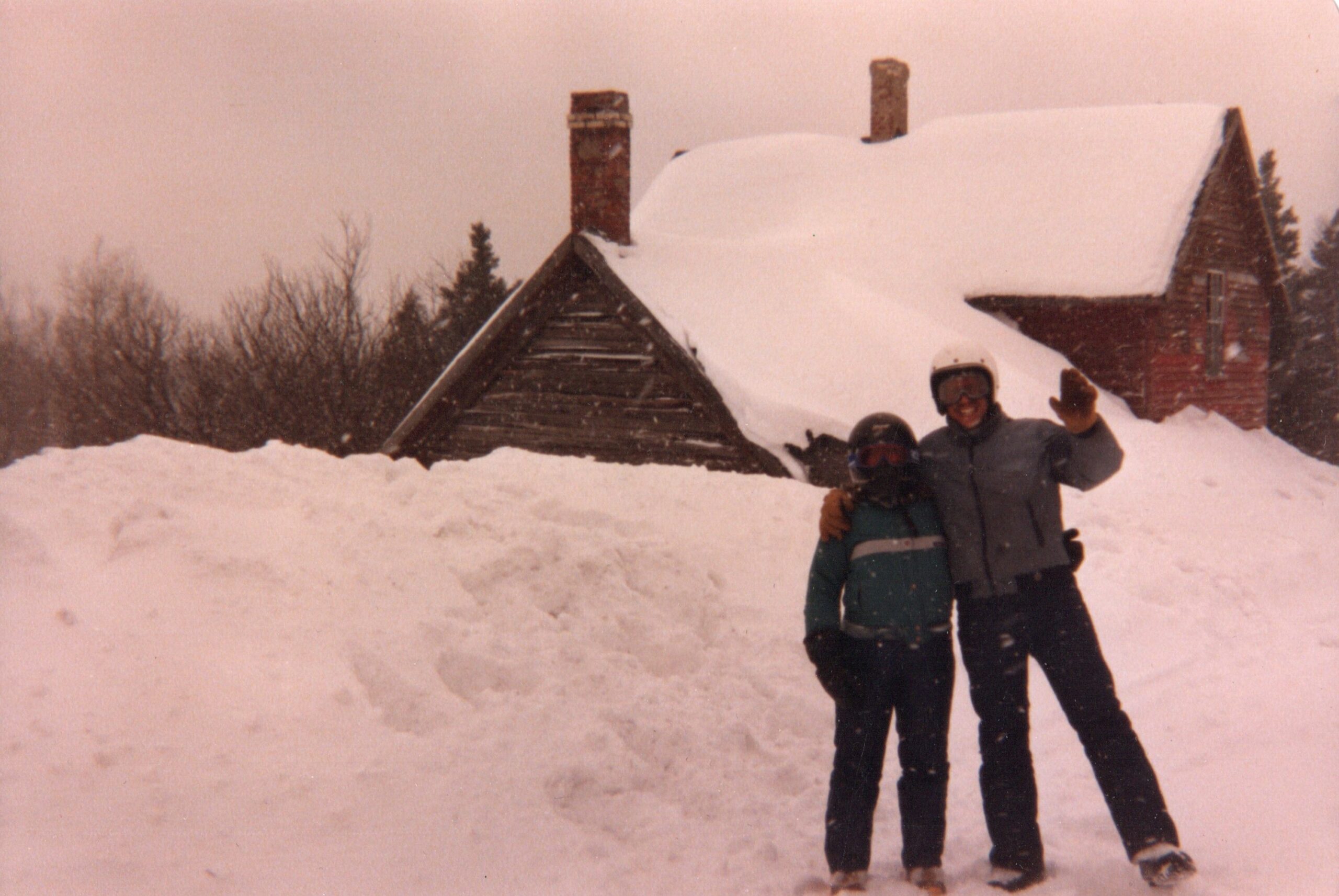 Two people in winter gear pose and wave from a snow-covered yard in front of a cabin.