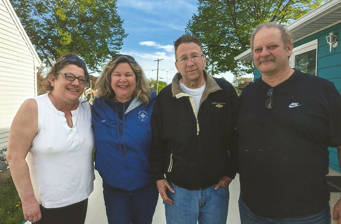 Four adults posing together on a sunny suburban street, smiling at the camera.