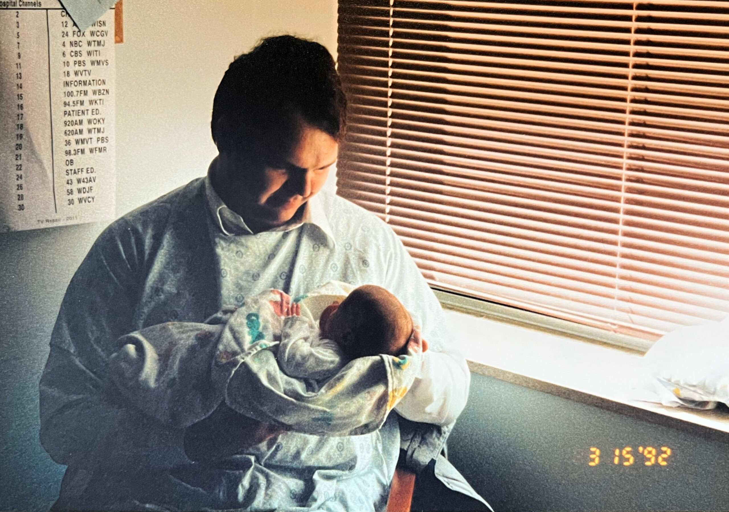 Man in a hospital gown sits holding a newborn baby in a ward with wooden blinds nearby, date stamp visible on screen edge.