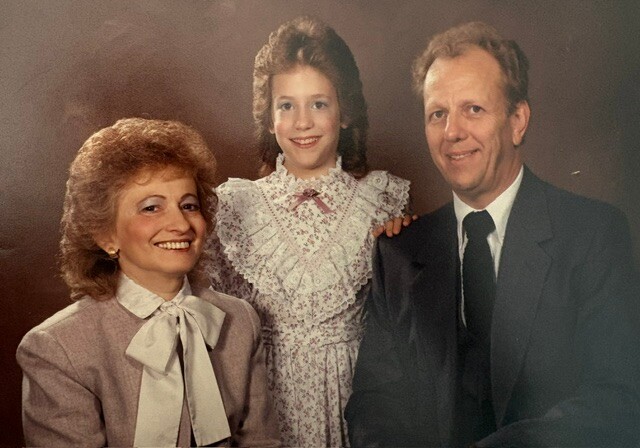 Three people posing for a family portrait: an older woman in a lavender suit, a girl in a lace dress, and a man in a dark suit, all smiling in the studio.