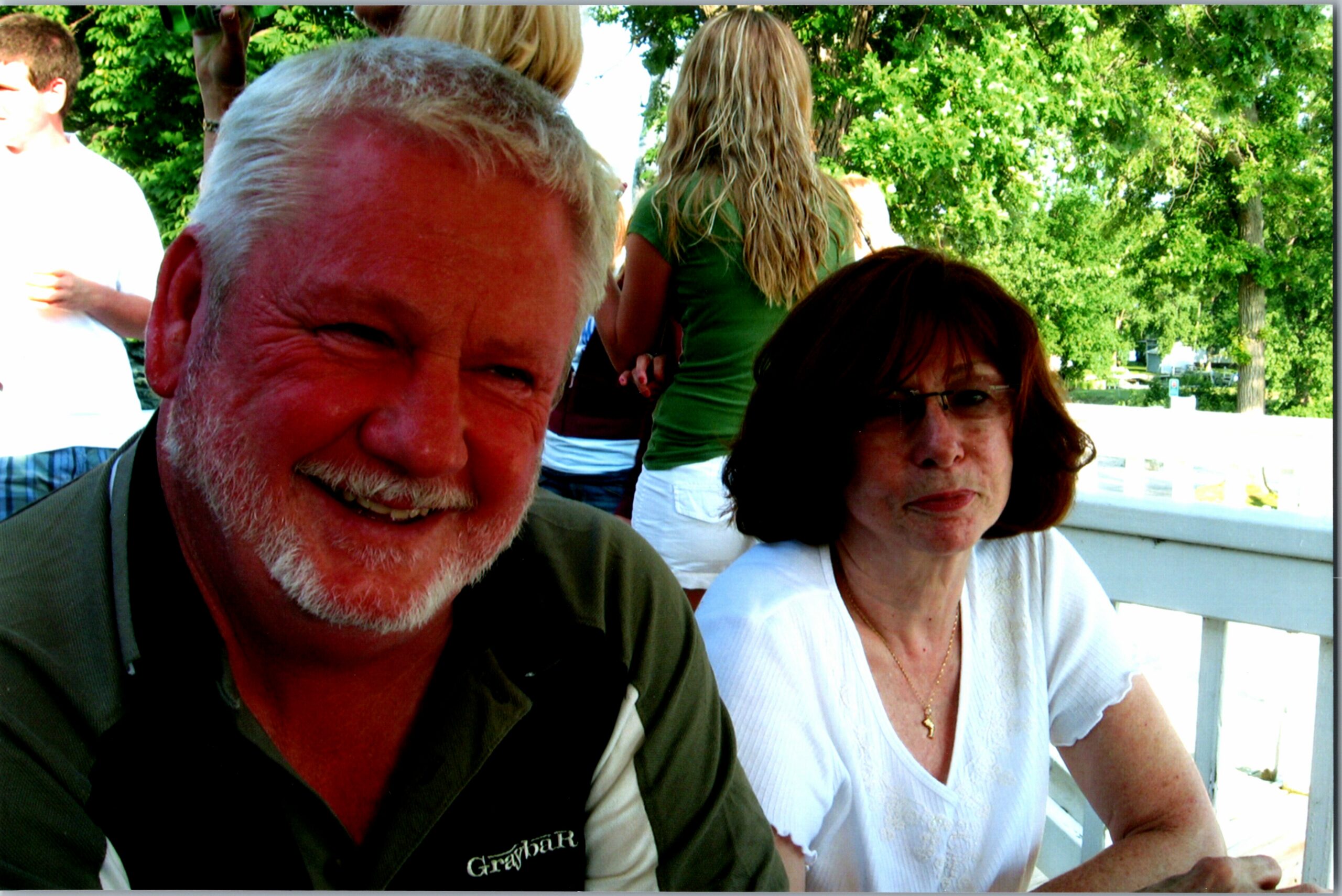 Two older adults posing for a selfie outdoors, man with gray beard in foreground and woman with short brown hair in a white blouse nearby, green trees behind them.