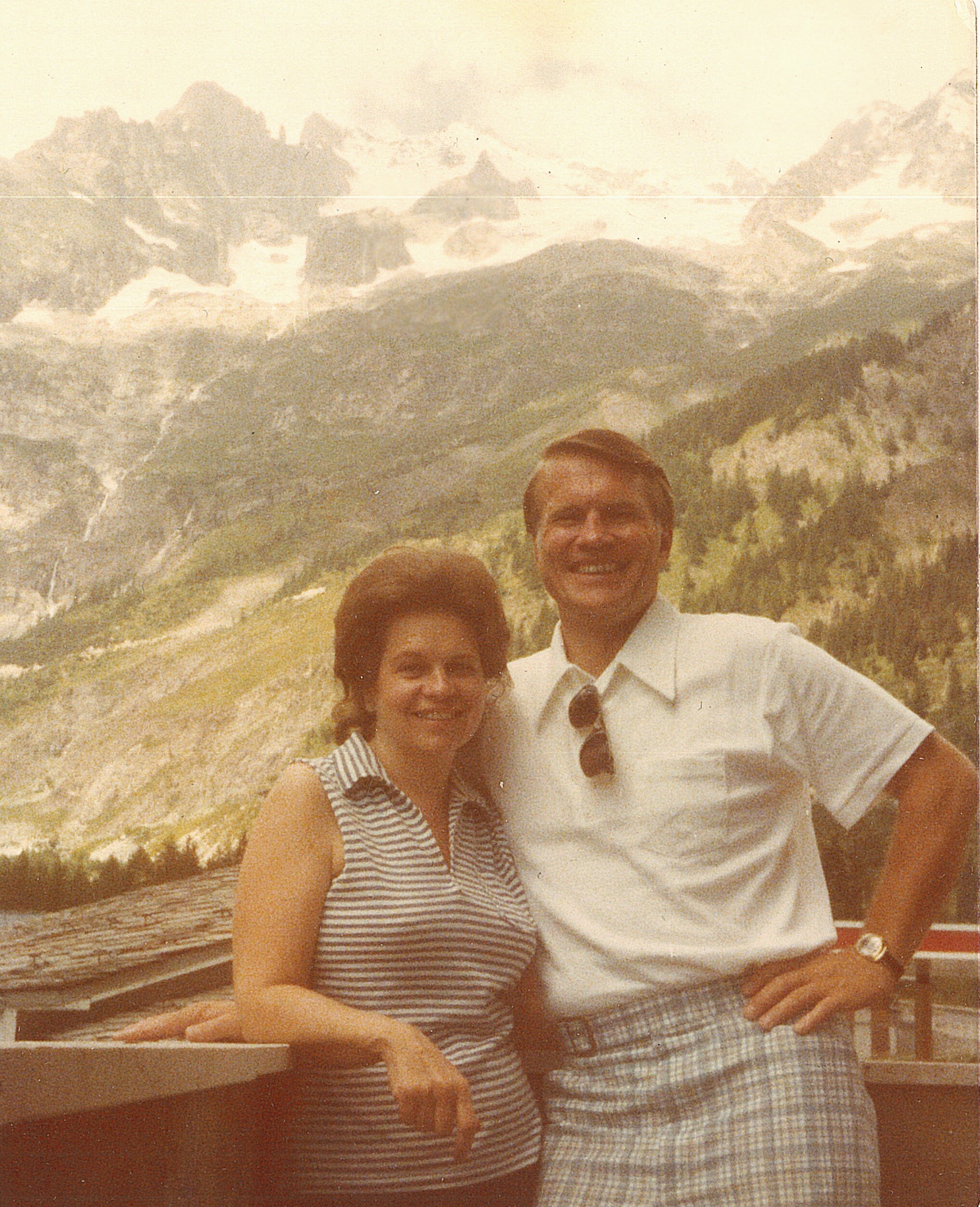 Smiling couple posing together on a railing with a rugged mountain range behind them.