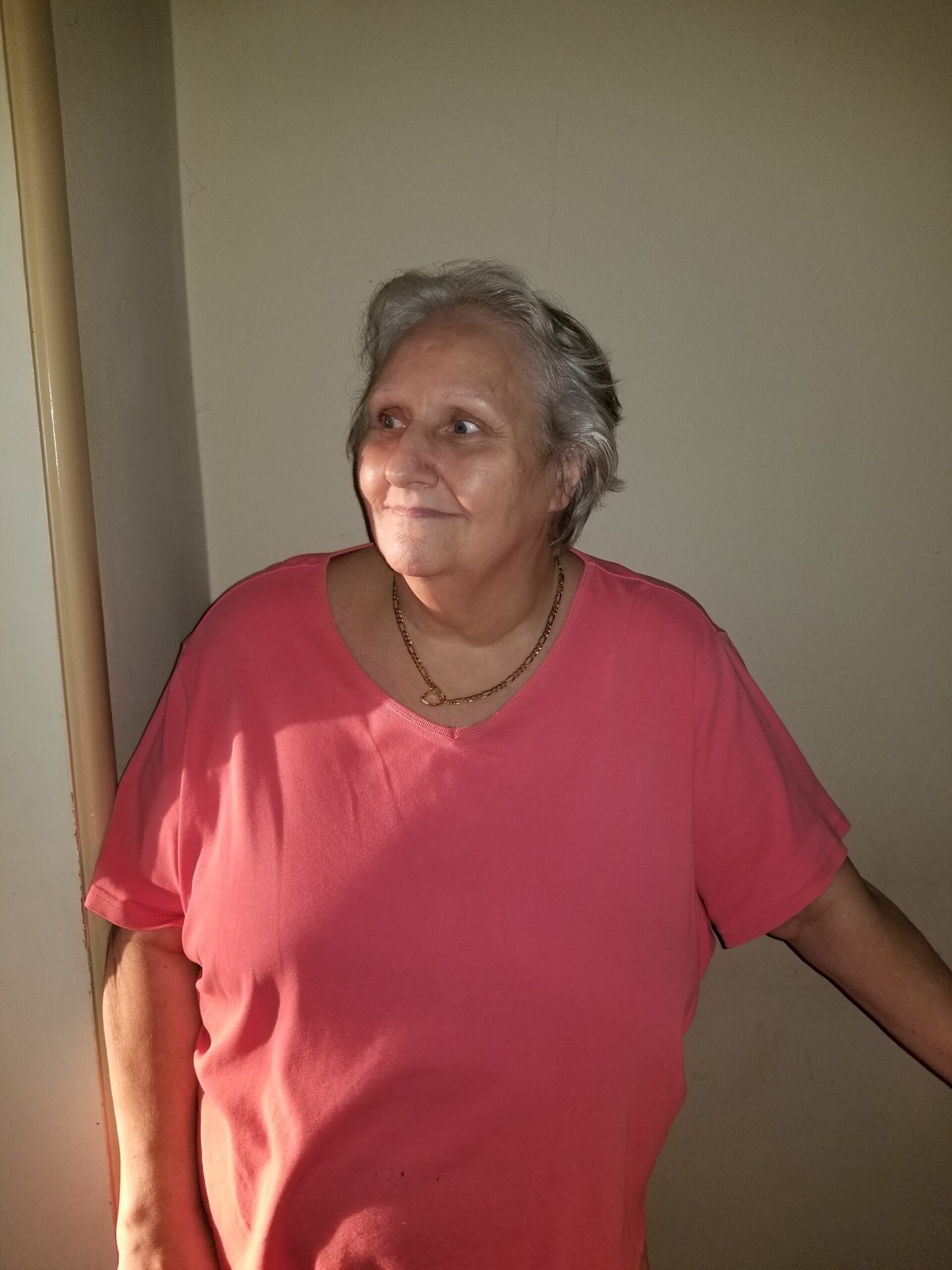 Older woman with short gray hair in a pink shirt, standing indoors by a door frame, looking to her left with a gentle smile.
