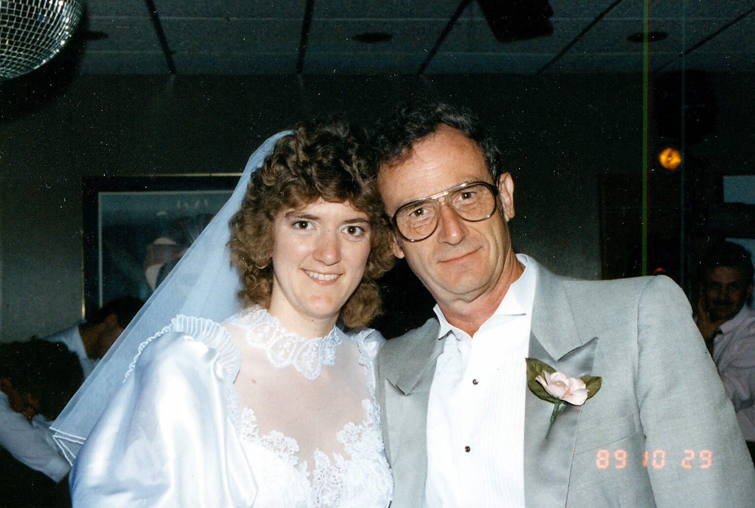 Bride in a white lace wedding dress and veil posing with a man in a light gray suit at a wedding reception, both smiling.