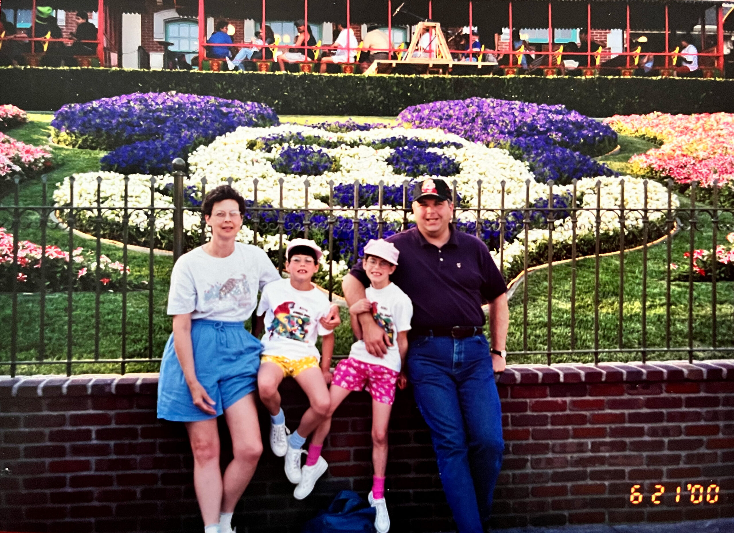 Family group posing in front of a circular flower bed with purple, white, and pink blooms and a brick wall behind them.