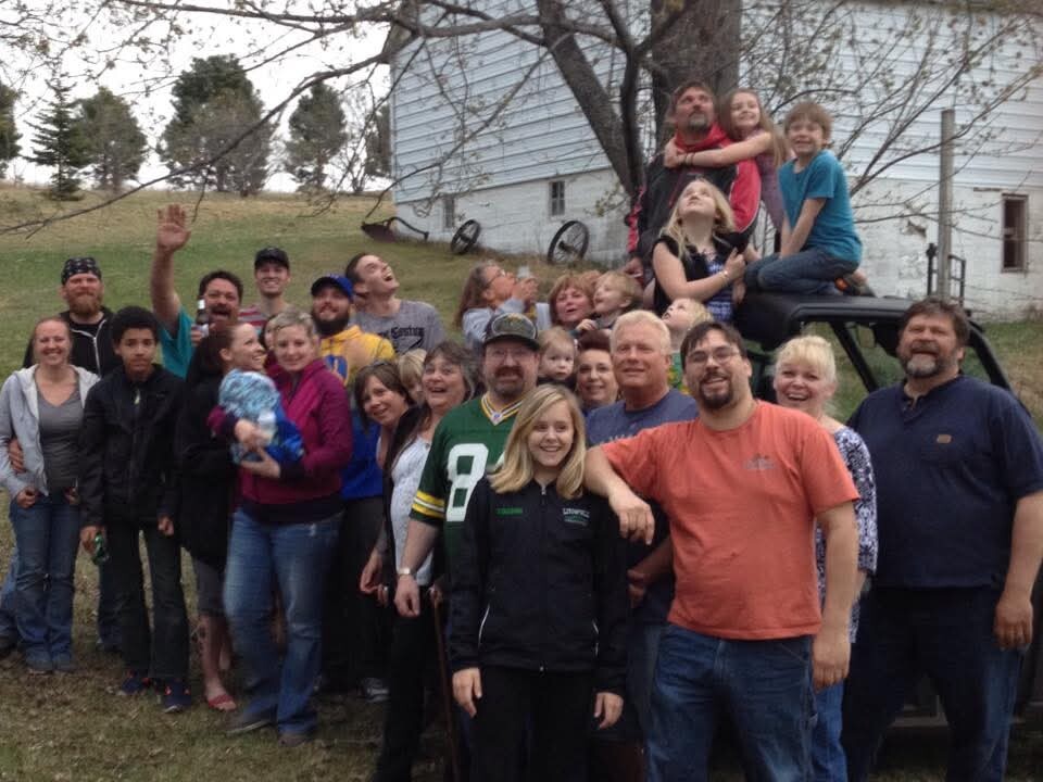 Large group of people posing together outdoors in a yard, with a white house in the background and some children sitting on a trailer.