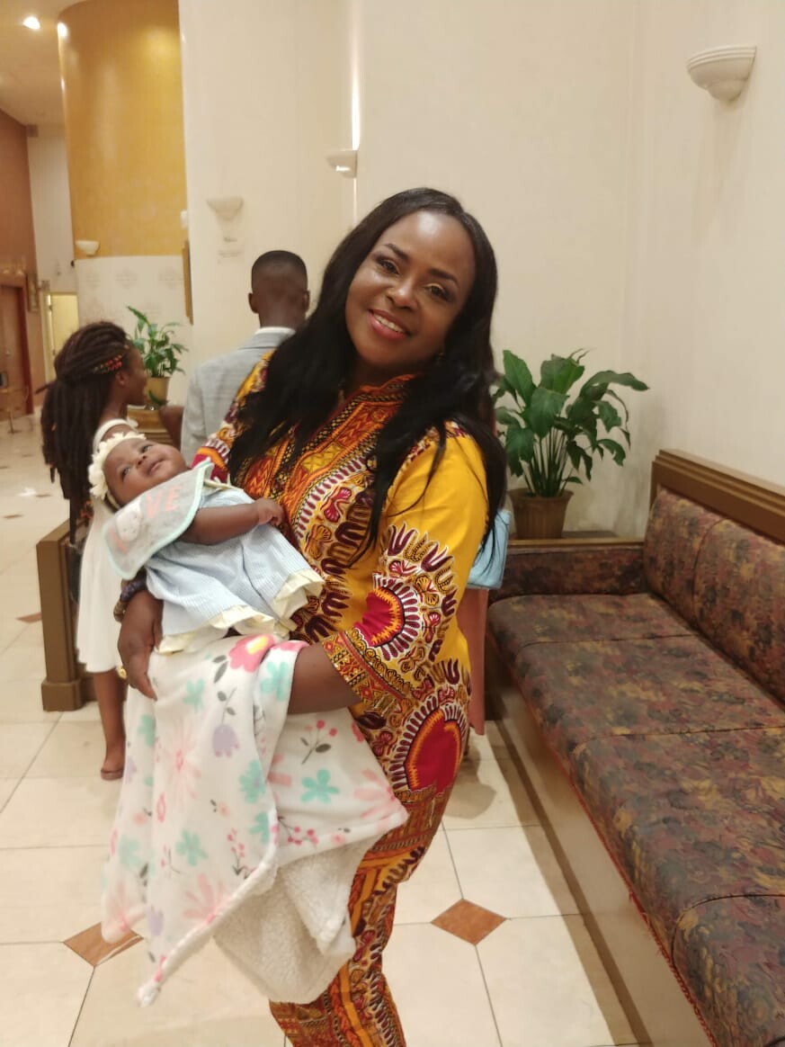 Smiling woman in a vibrant yellow African-print dress holds a baby wrapped in a pastel floral blanket in a hotel lobby.