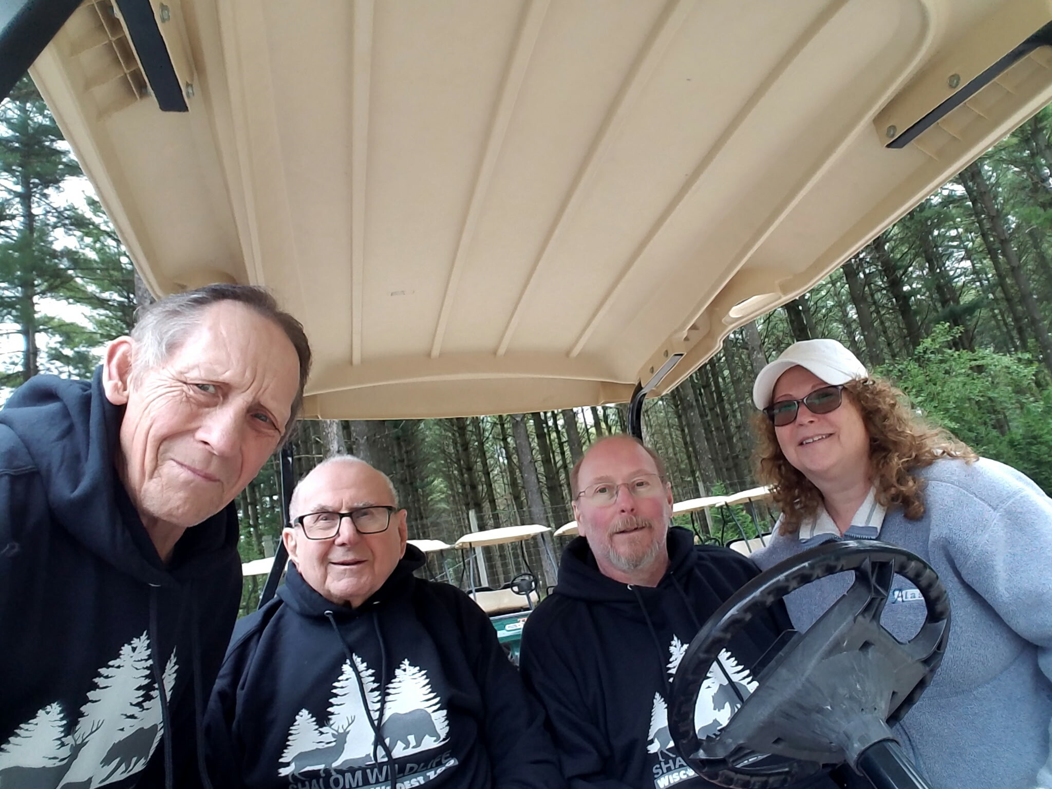 Four people taking a selfie in a golf cart outdoors, wearing matching black hoodies with white tree silhouettes; a fence and trees in the background.