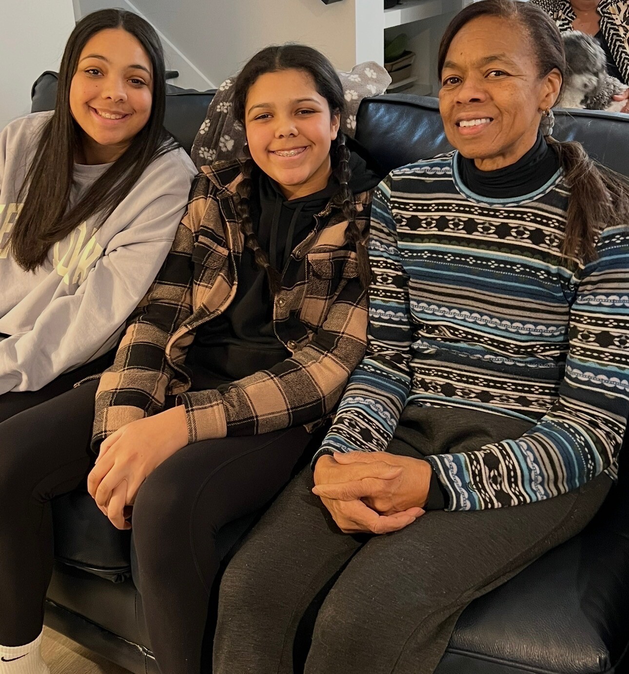 Three women of different ages sitting on a black leather couch, smiling at the camera.
