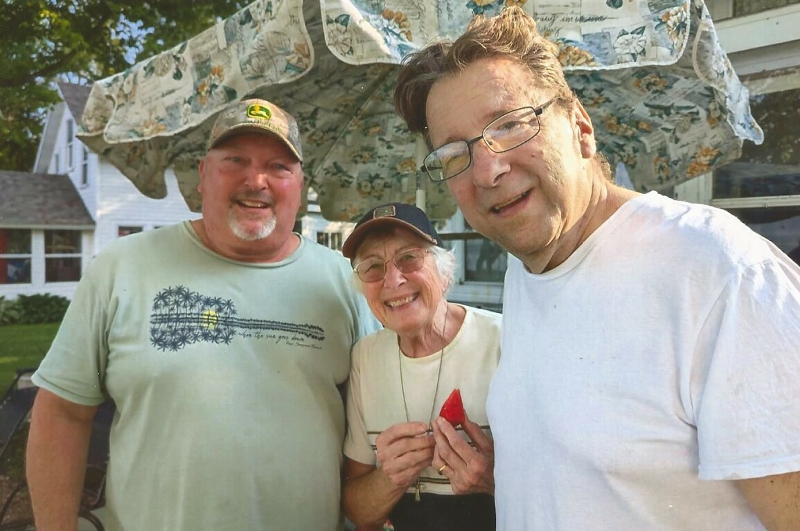 Three smiling adults posing in a backyard on a sunny day under a patterned canopy (left man in a green shirt, center woman with cap, right man in white T-shirt).