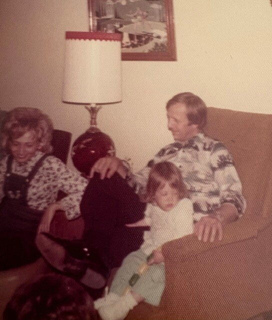 Family scene in a living room: woman on the left, man on the right with a child on his lap, seated on a brown sofa with a lamp and painting in the background.