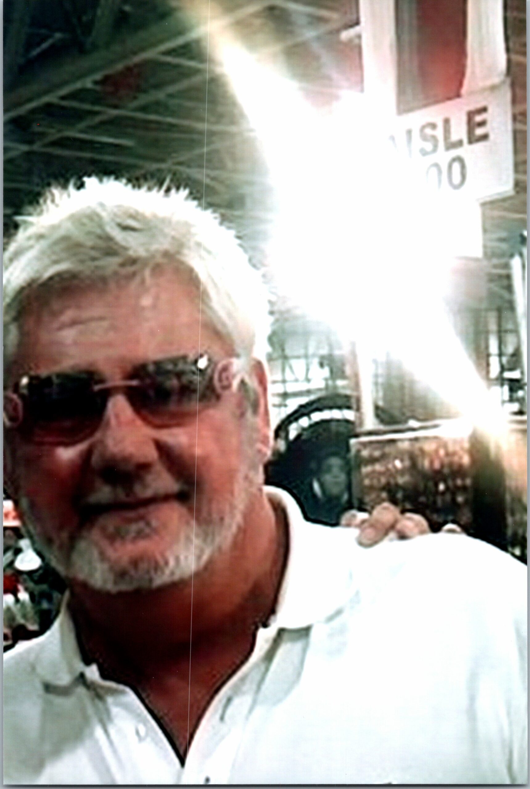 Close-up selfie of a man with white hair and sunglasses, outdoors with a storefront sign in the background.