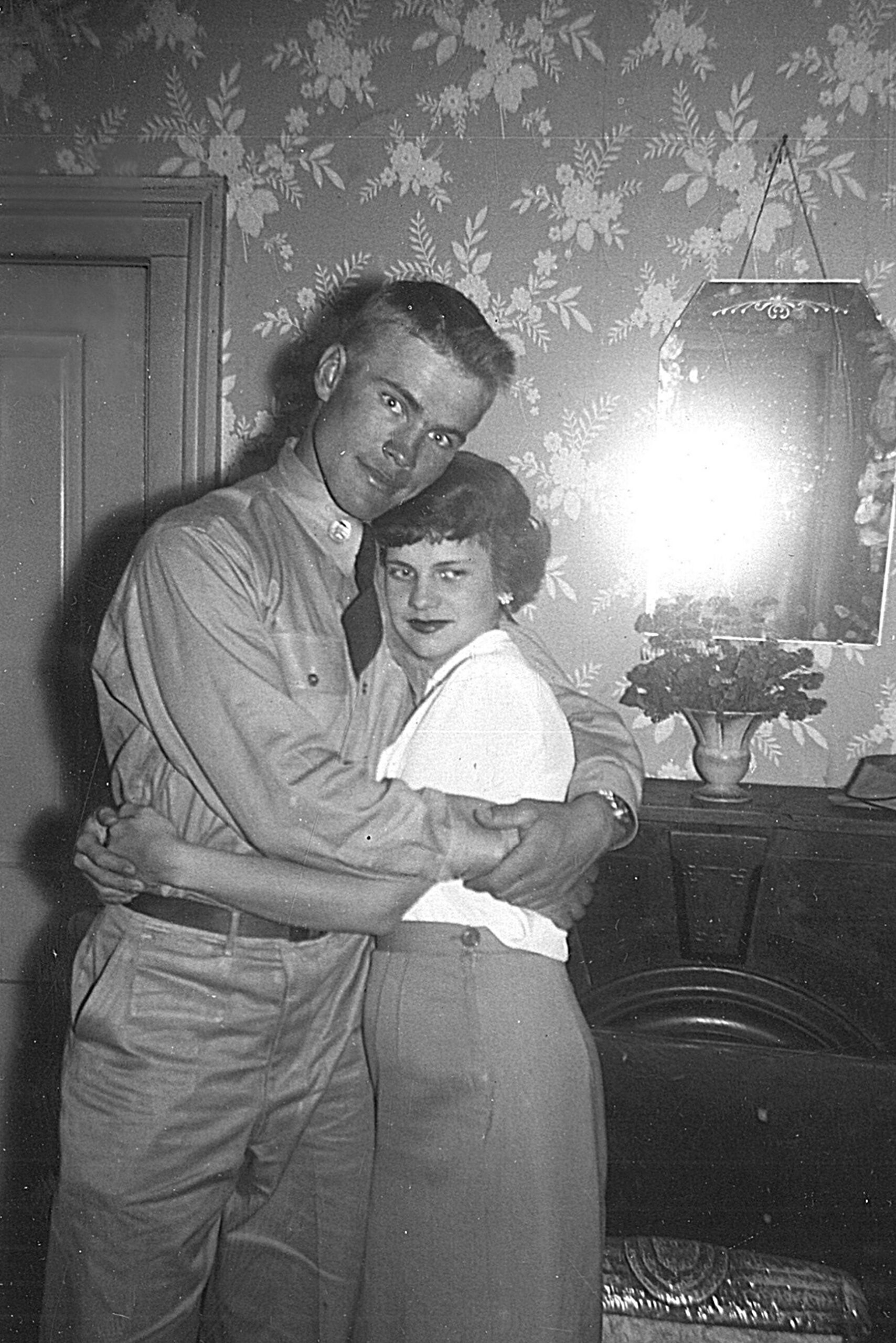 A couple hugs and smiles at the camera in a room with floral wallpaper and a lamp on a table behind them.