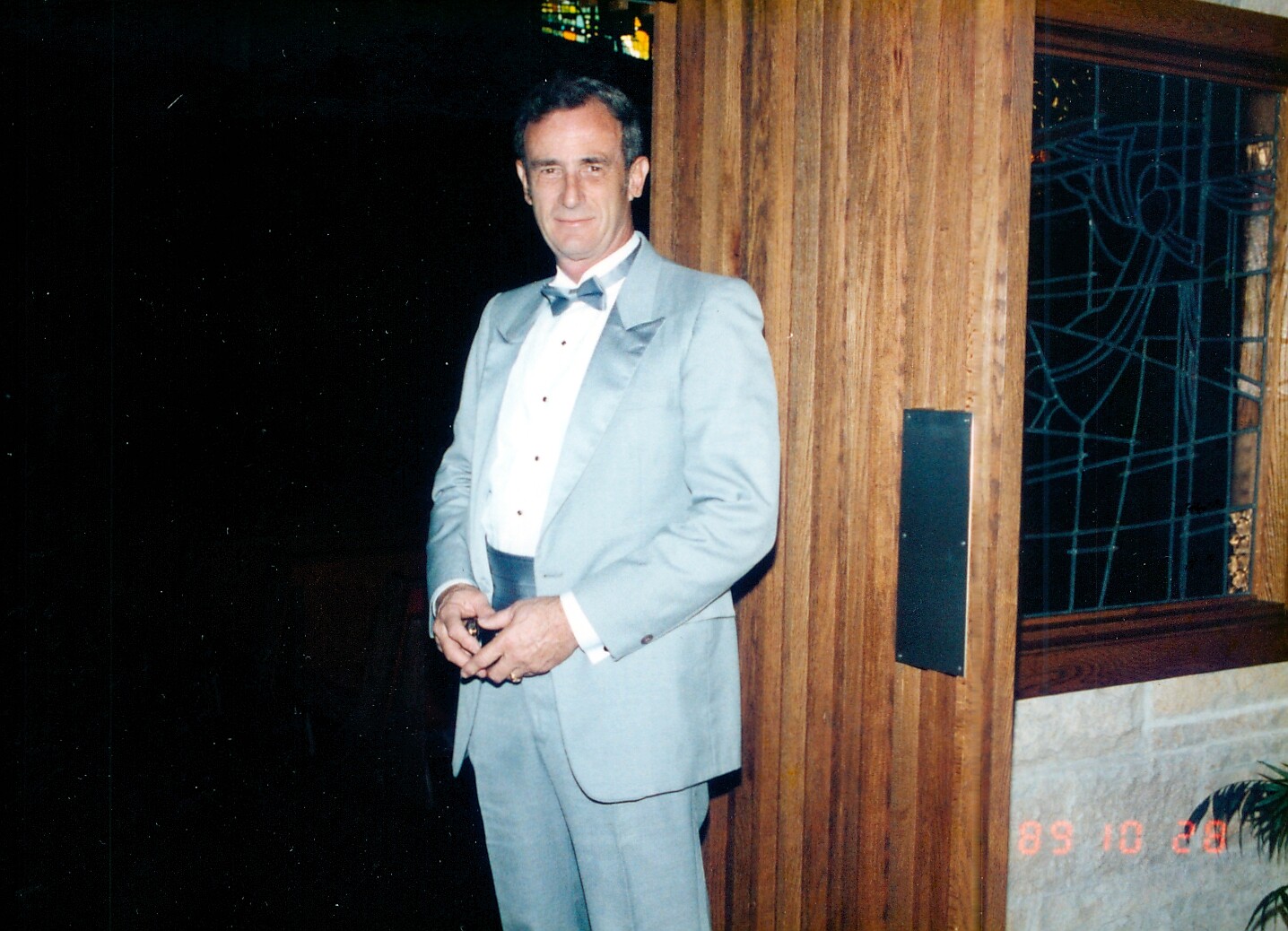 Man in a light blue tuxedo and bow tie standing by a wooden wall at night