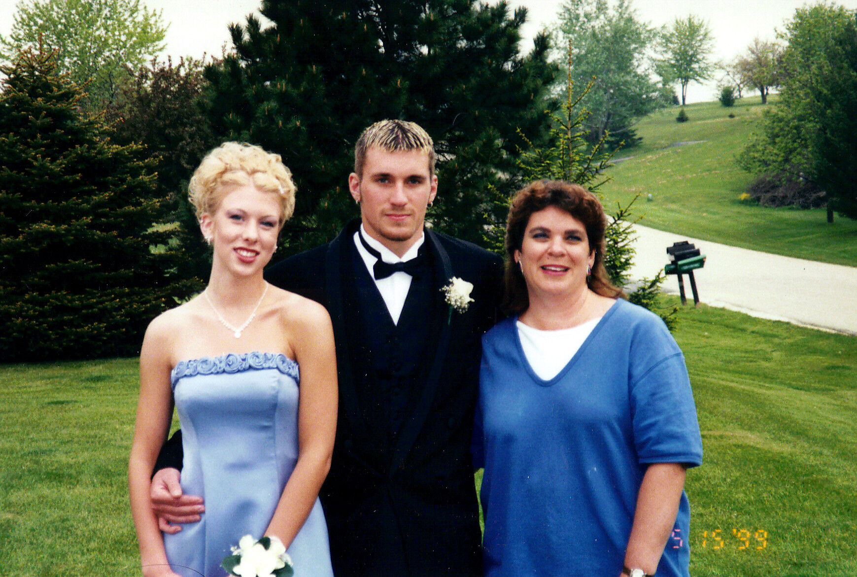 Three people pose outdoors on a grassy area: a young woman in a light blue strapless dress, a man in a black tuxedo, and another woman in a blue top, all smiling.