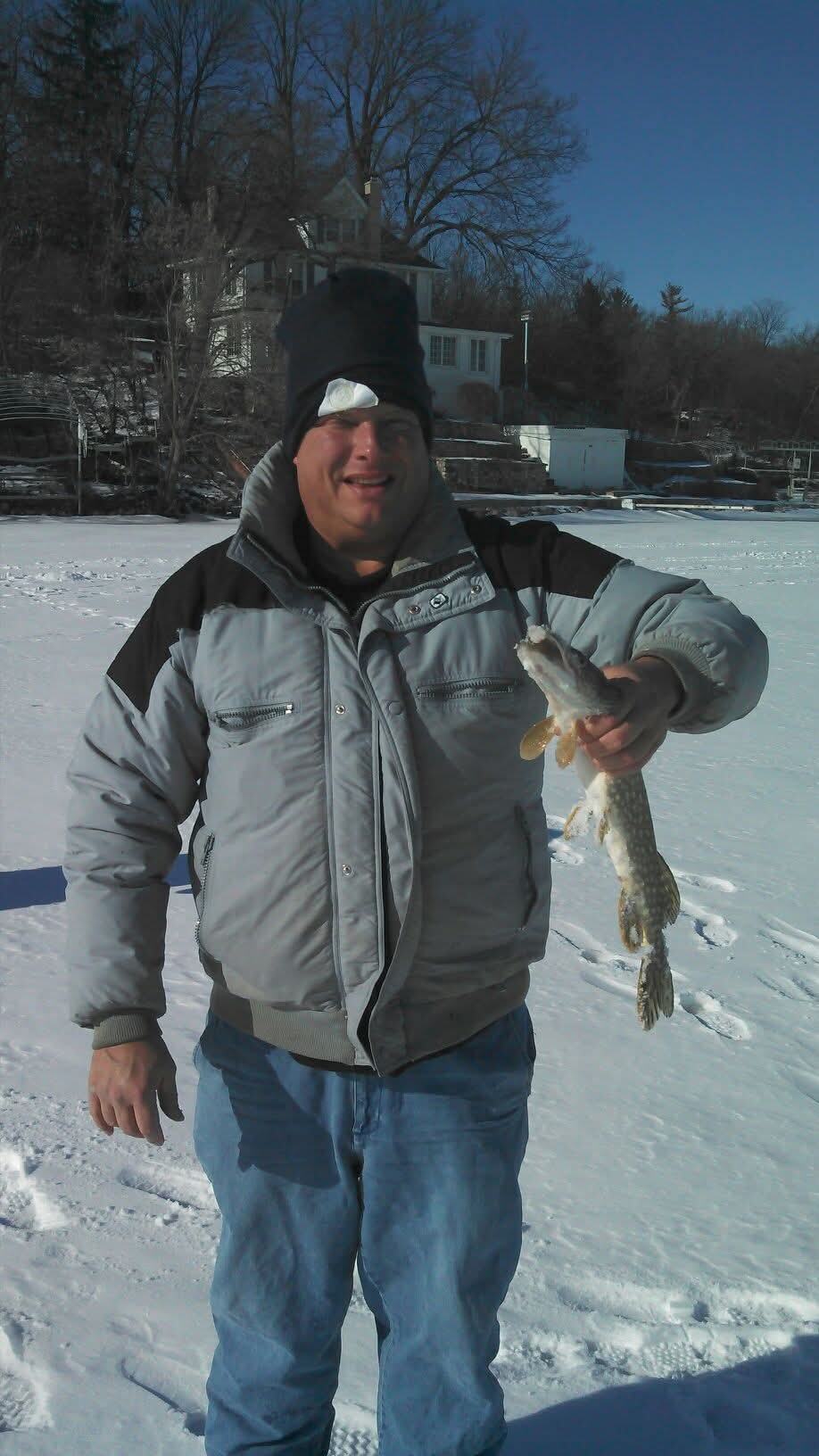 Man in a gray winter jacket and black beanie on a snowy shoreline holding a long, snouted fish.