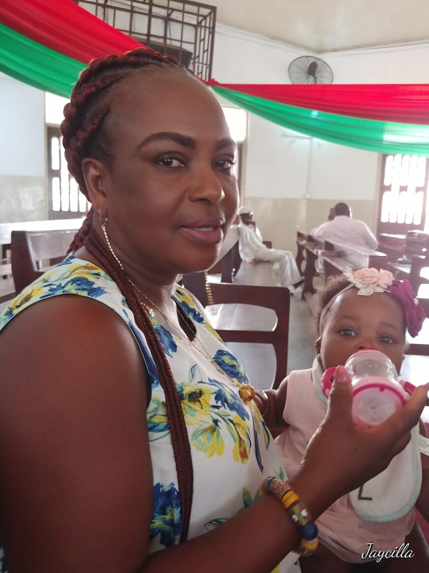 Mother with braided hair feeding a baby with a bottle in a hall decorated with red and green fabric.