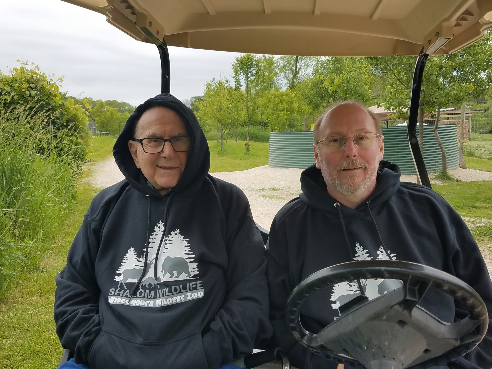 Two men in a golf cart outdoors, wearing black hoodies with white tree graphics, smiling at the camera.