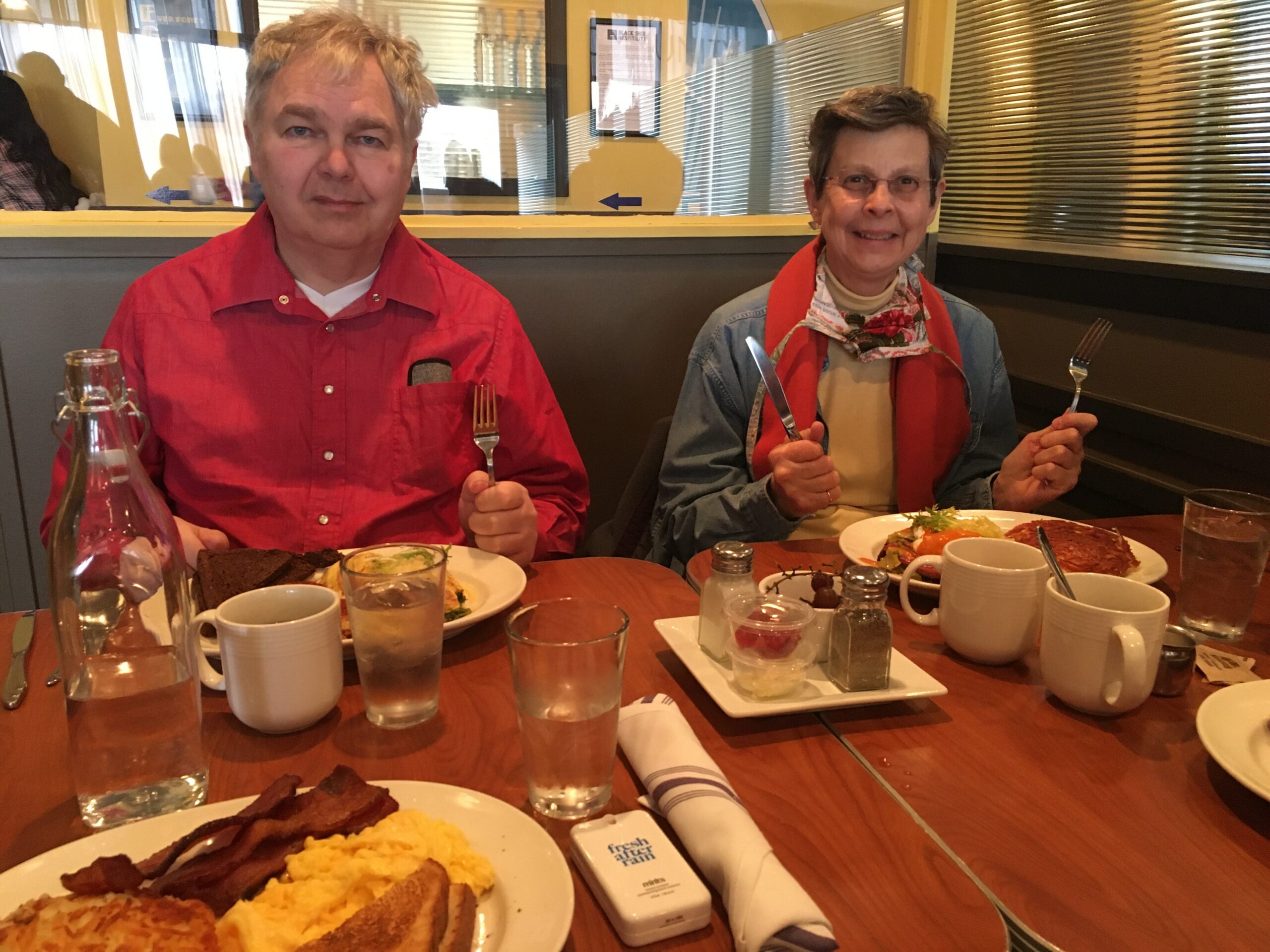 Two people sit at a table in a casual restaurant, enjoying a meal together with plates of food and drinks in front of them.