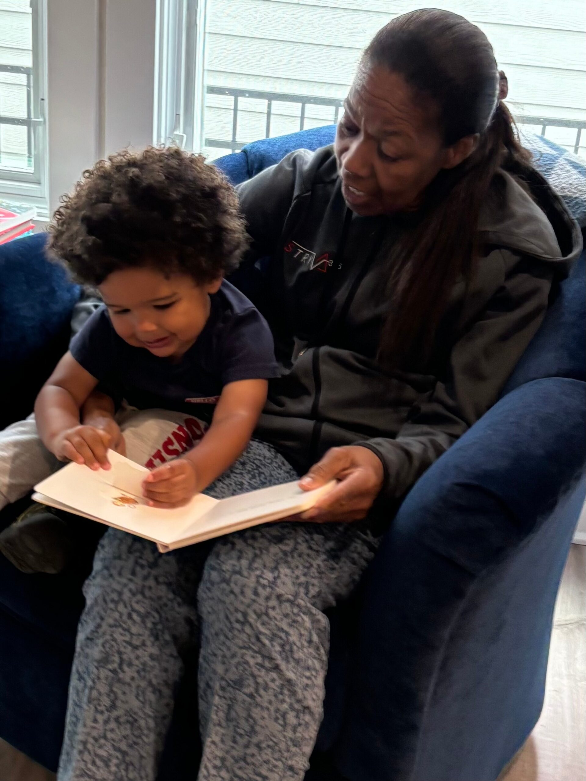 A woman sits on a blue chair reading a children’s book with a young child on her lap.