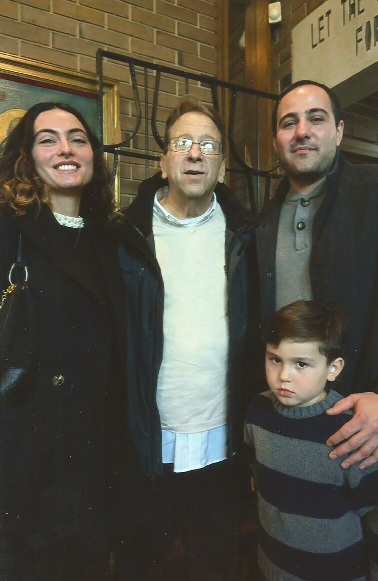 Four people posing indoors against a brick wall: woman on the left, an older man with glasses in the middle, a man on the right, and a young boy in front wearing a striped sweater.