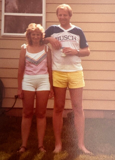 Two adults posing outside a beige house: a woman in a pink top and white shorts, and a man in a Busch shirt with yellow shorts, holding a drink.