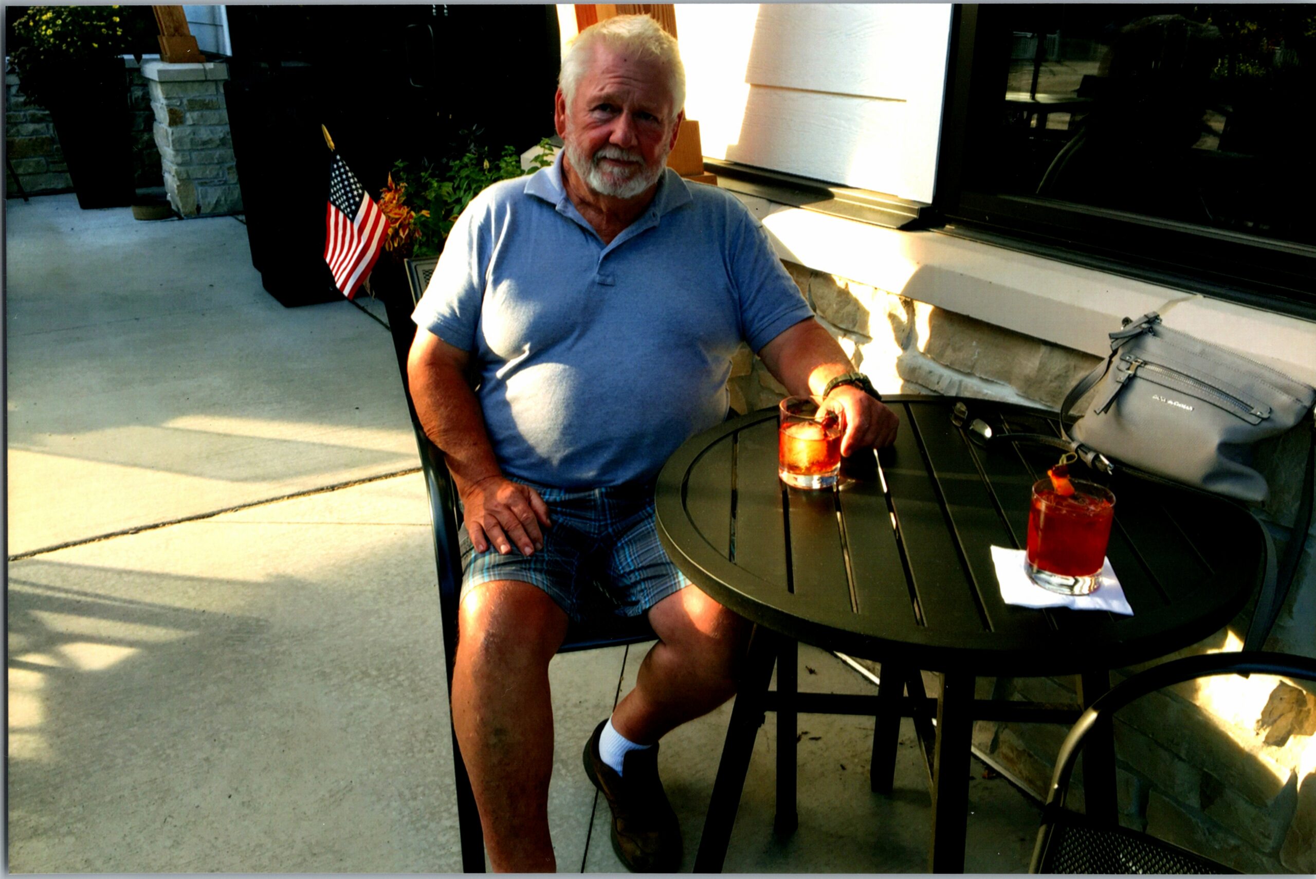 Older man with white beard sits at a dark patio table outdoors, holding a iced drink in a short glass, wearing a light blue polo and plaid shorts, with an American flag in the background.