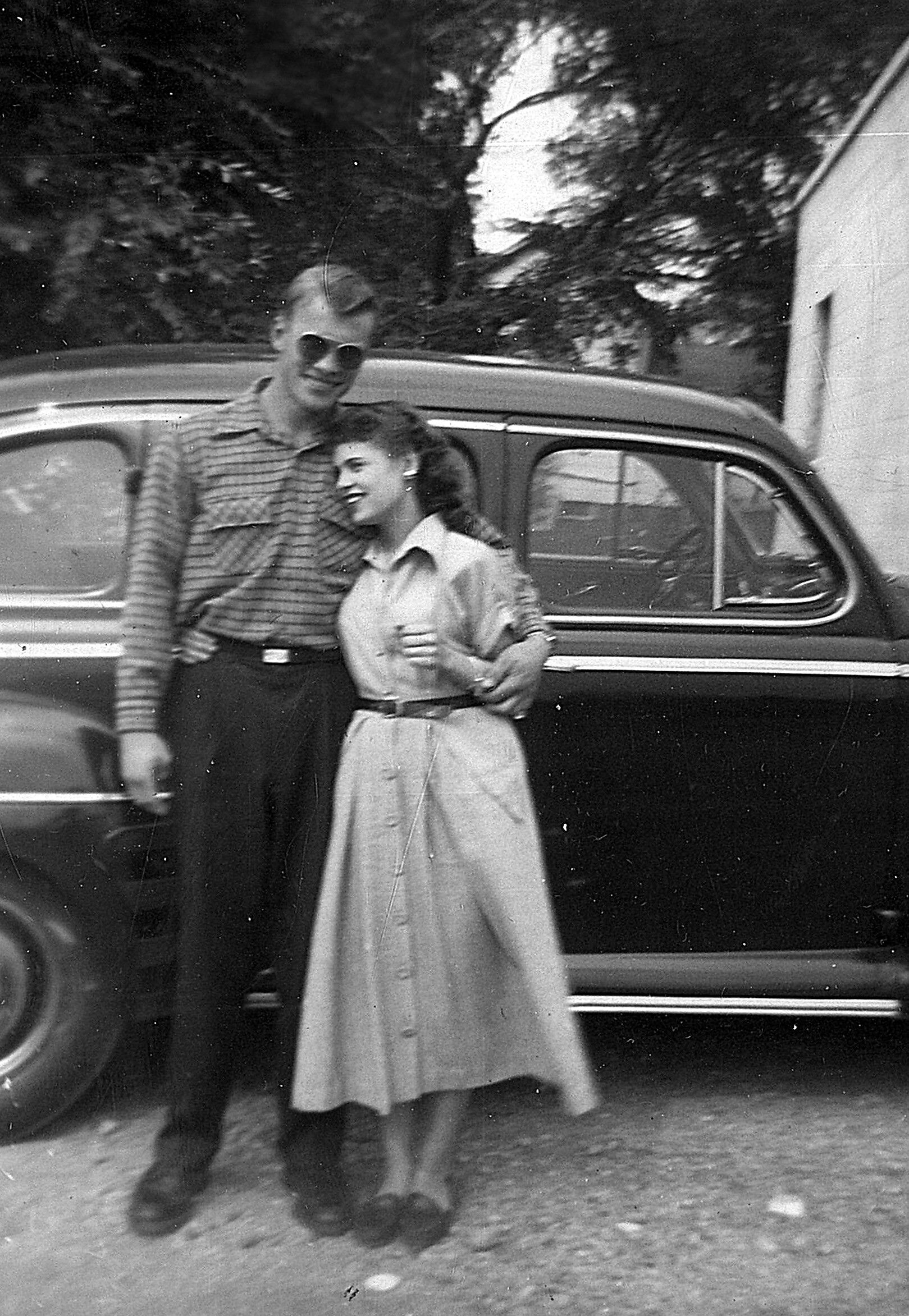 Couple posing arm in arm beside a vintage car, smiling at the camera.