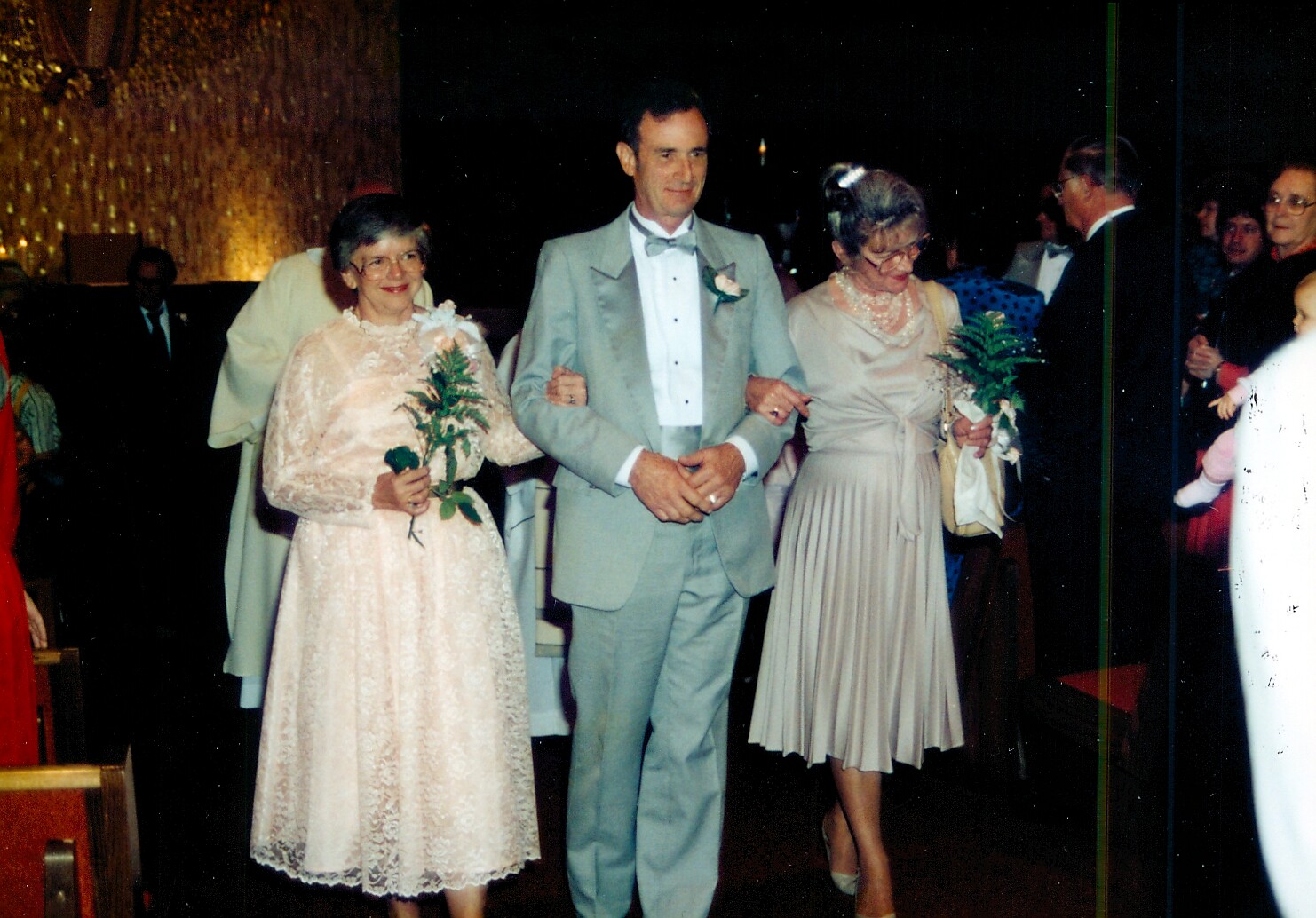 Man in a light gray suit walks between two women in pastel dresses at a formal event, each holding a small bouquet or fern.