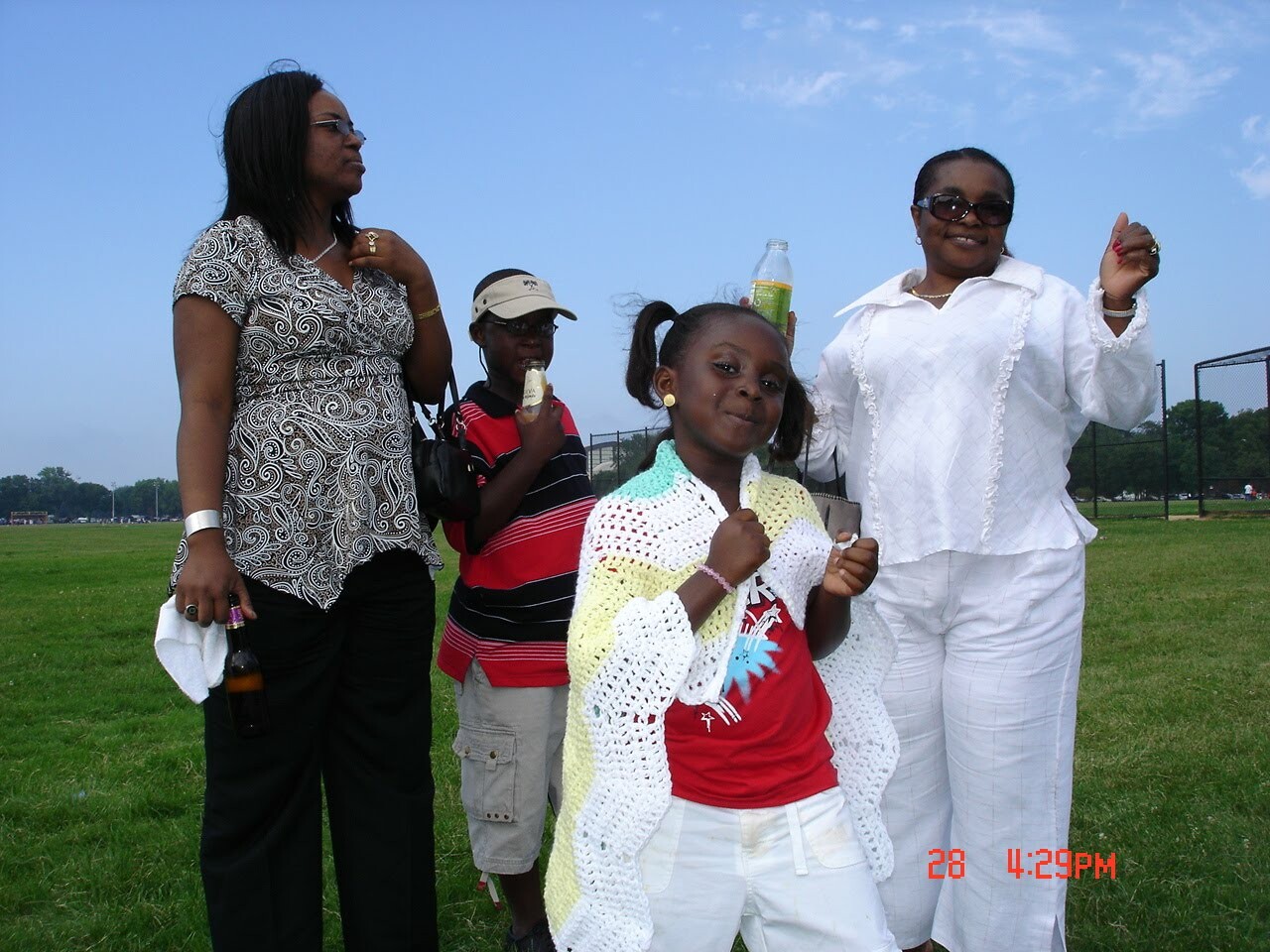 A group of four people posing on a sunny park field: a young girl in front wearing a white crocheted cardigan over a red shirt, flanked by adults holding drinks behind her.