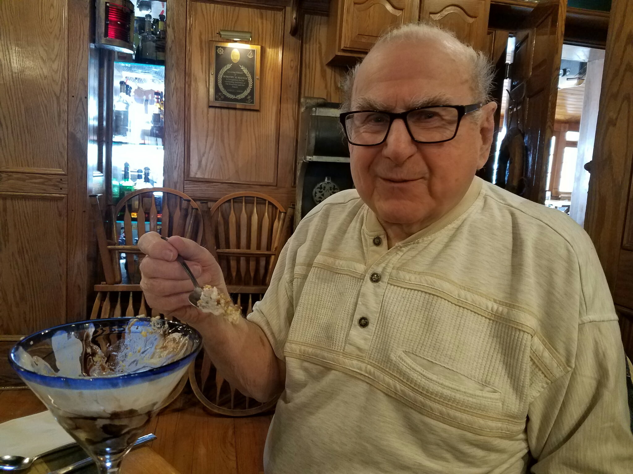 Older man with glasses sits at a wood-paneled restaurant table, holding a spoon over a partially eaten ice cream bowl in front of him.