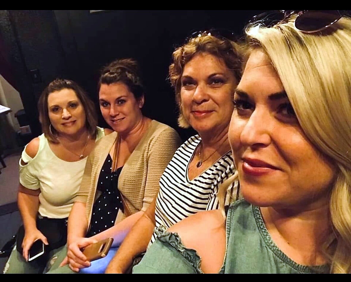 Four women sitting closely together, taking a selfie in a dim indoor space, all smiling at the camera.