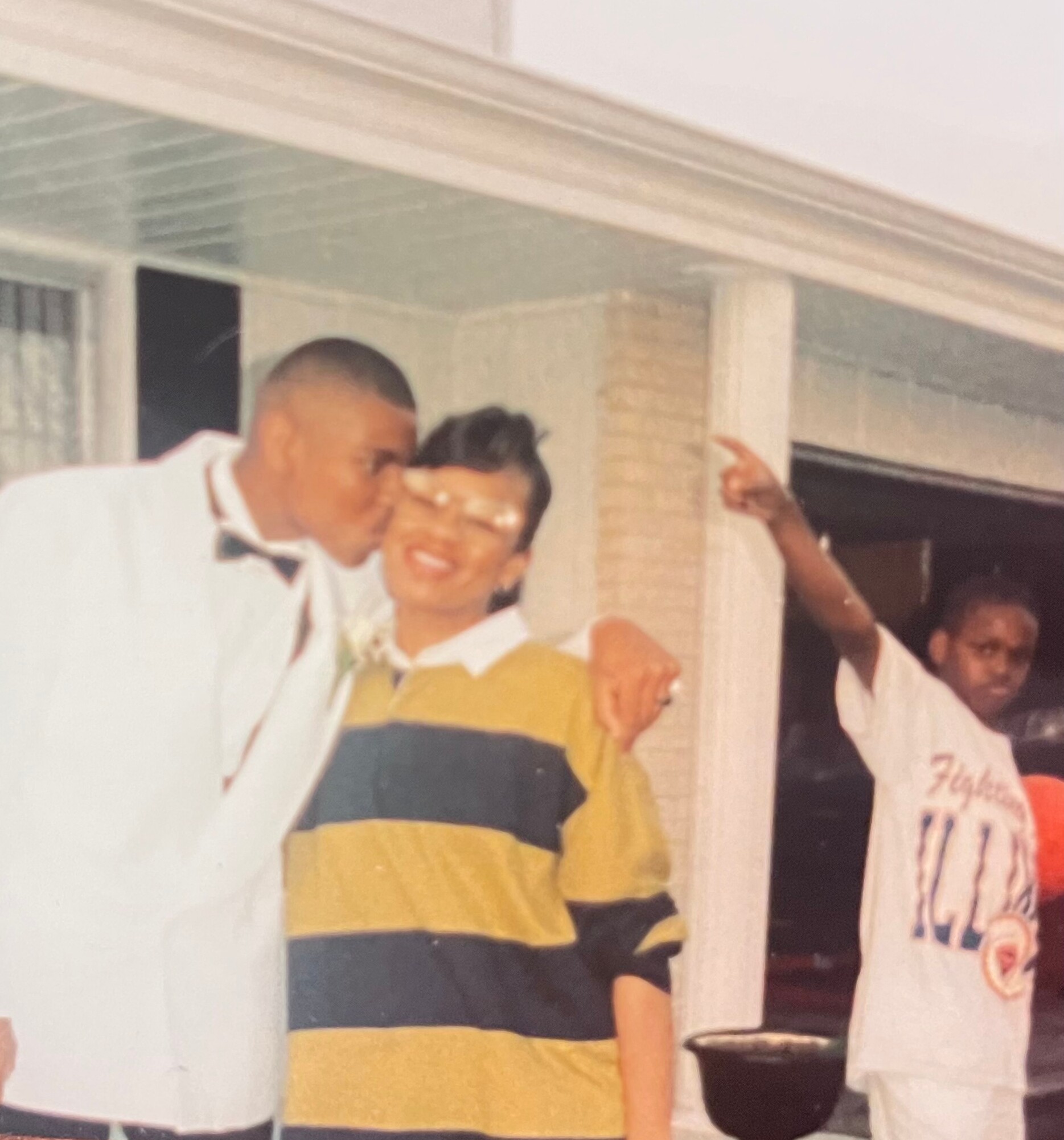 Two men in the foreground hug and smile at a porch party, with guests visible in the background.