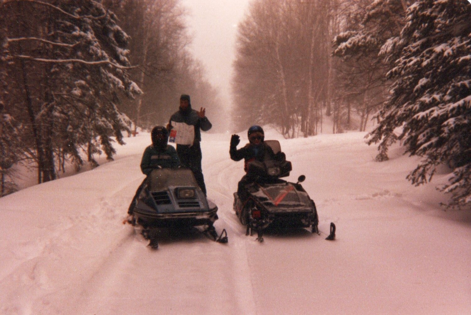 Three people in winter gear pose with two snowmobiles on a snowy forest trail; one stands waving with a sign while the others sit on their machines.