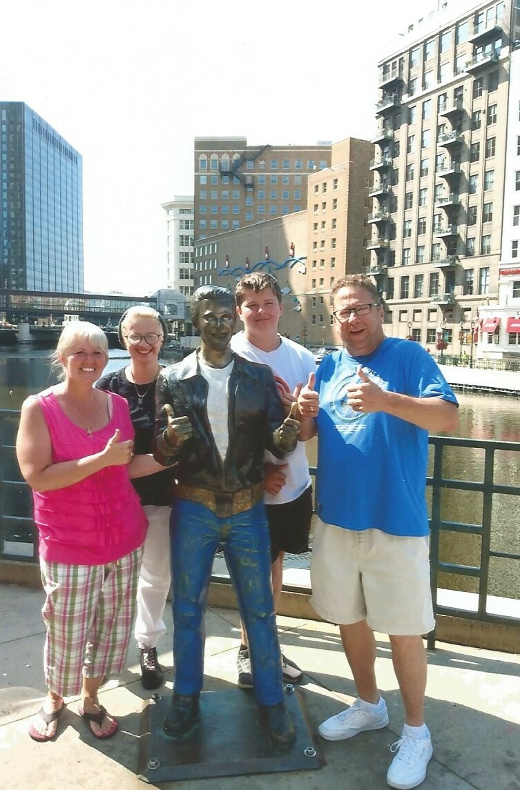 Four smiling adults pose with a life‑size bronze-like statue of a man in a leather jacket on a city waterfront promenade.