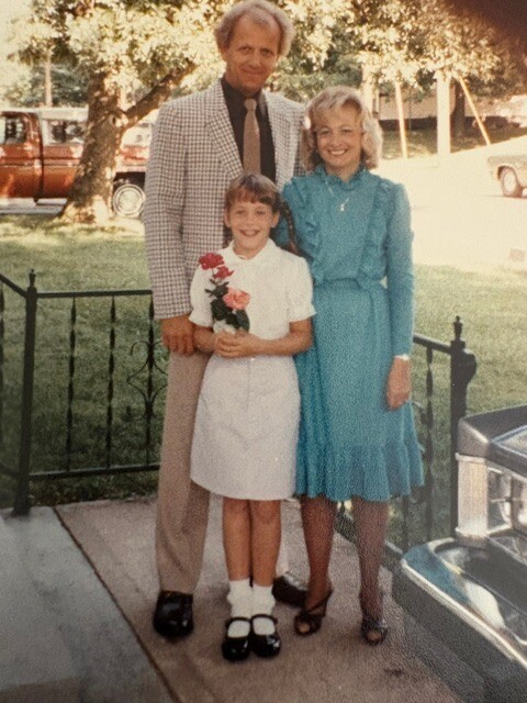 Family of three posing outdoors: father in a plaid blazer, mother in a teal dress, daughter in a white dress holding flowers.