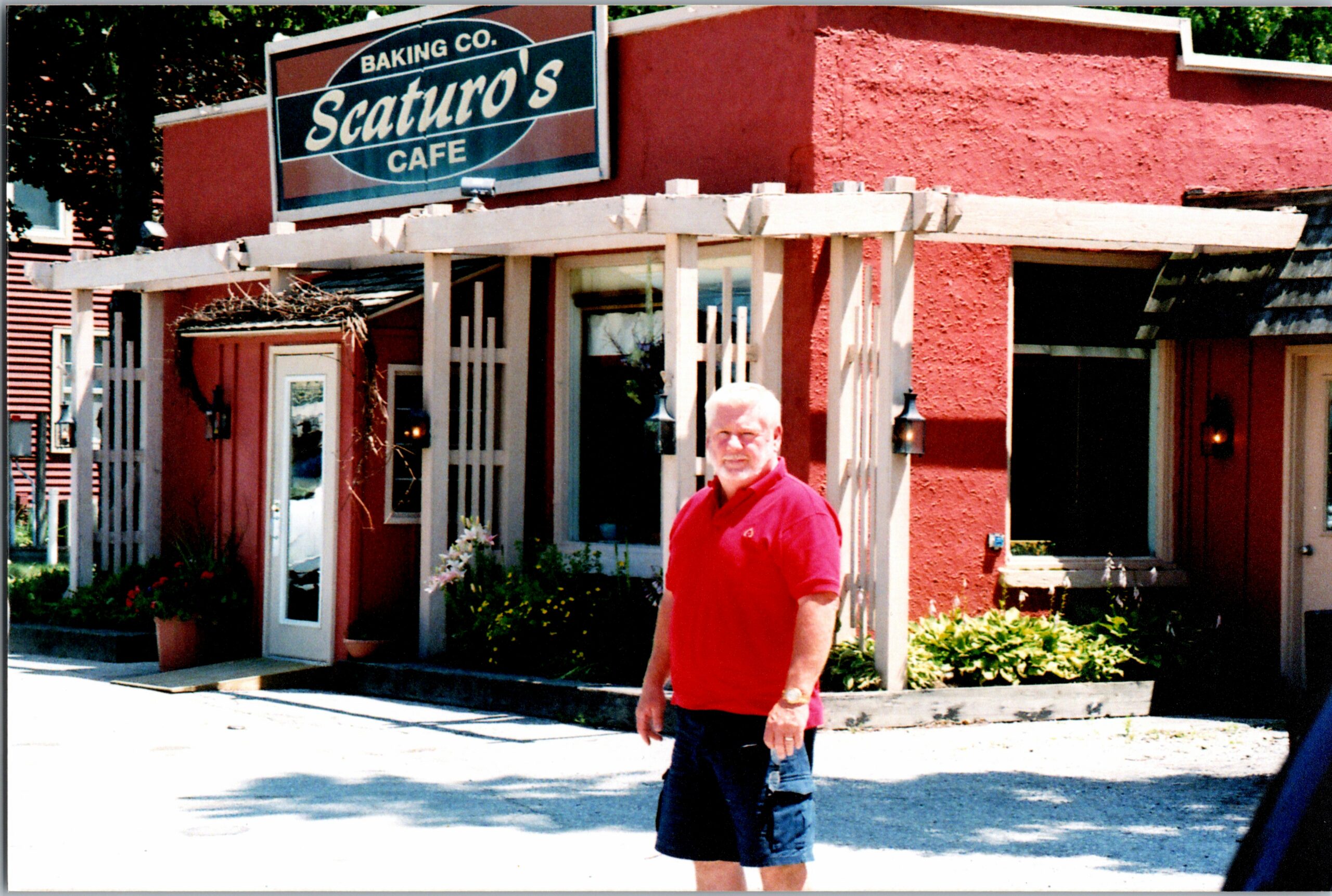 Man in a red polo standing outside Scaturo's Café, a red stucco building with a white pergola.