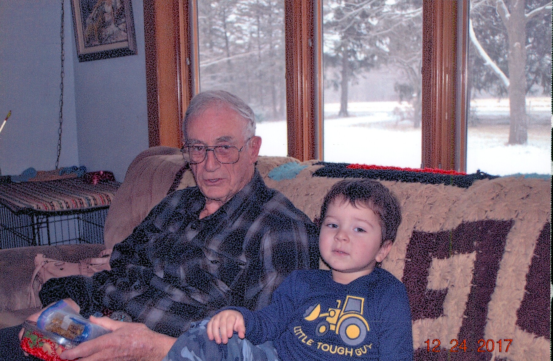 An elderly man with glasses sits on a couch next to a young boy, indoors by a window with a snowy yard outside.