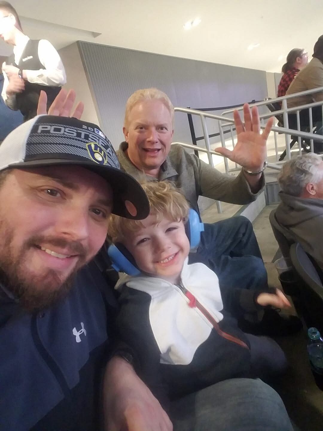 Three people sit in stadium seats taking a selfie: a man in a cap and beard, a smiling boy with blue earmuffs, and a man in the background waving hello.