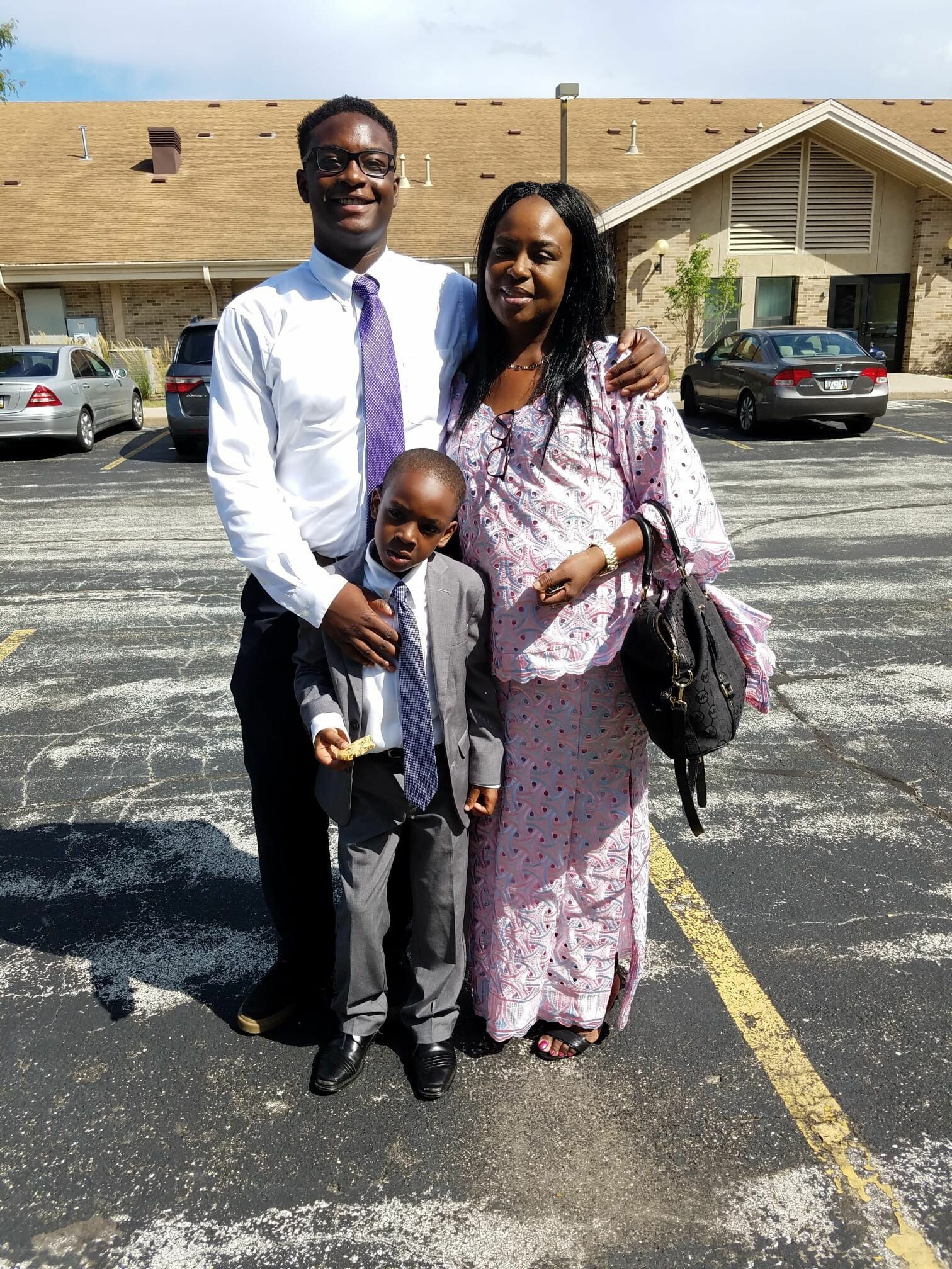 Three relatives posing together outside in a parking lot: a tall man in a white shirt and purple tie, a woman in a pink patterned dress, and a young boy in a gray suit.
