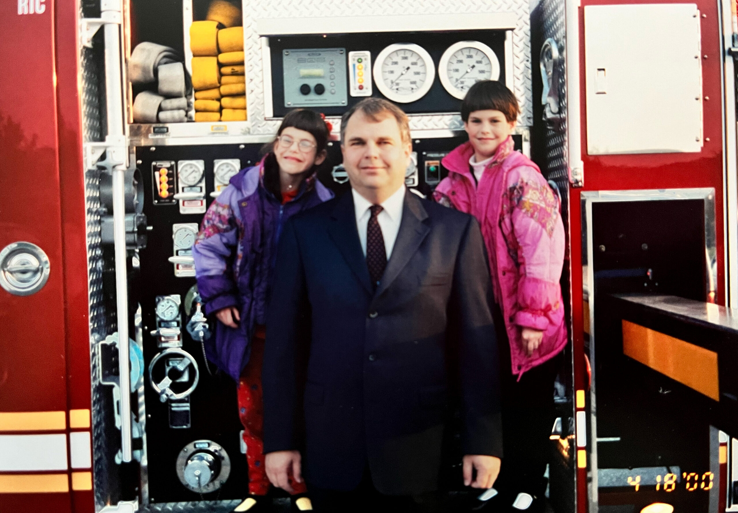 Adult man in a suit stands between two young girls in colorful jackets inside a red fire truck, posing for a photo with gauges and equipment behind them.