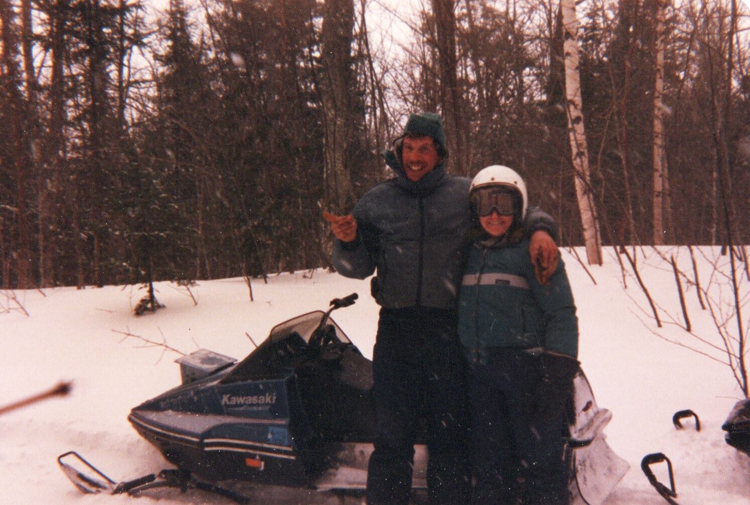 Two people in winter jackets and helmets pose with a blue Kawasaki snowmobile in a snowy forest.