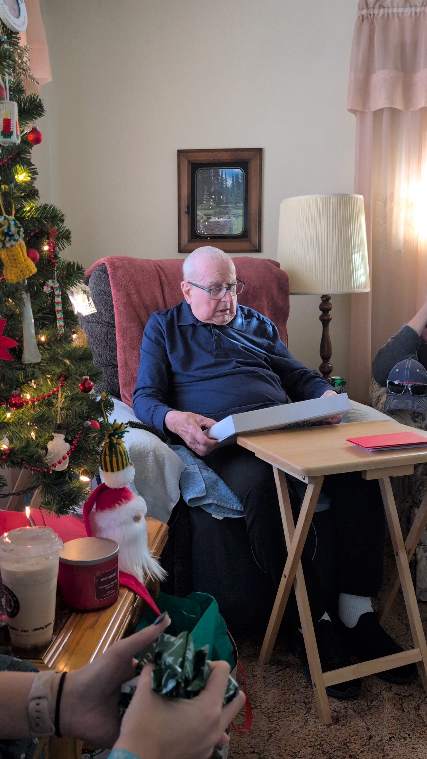 Elderly man in a blue shirt sits in a red armchair by a decorated Christmas tree, holding an open gift box.