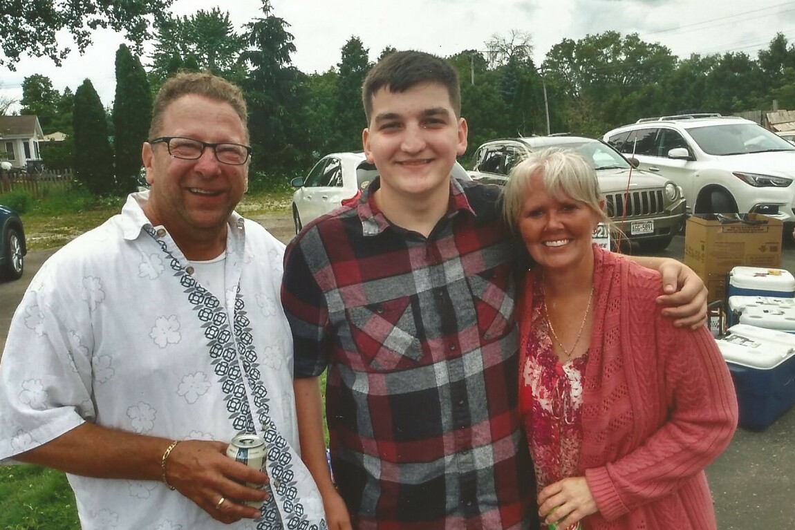 Three smiling people pose together outdoors at a casual gathering, with cars and coolers in the background.