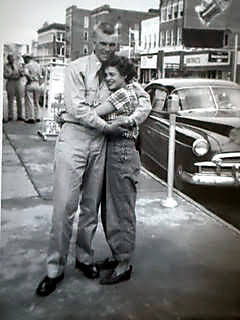 A couple dances closely on a city sidewalk beside a vintage car, with storefronts and bystanders in the background (black-and-white photo).