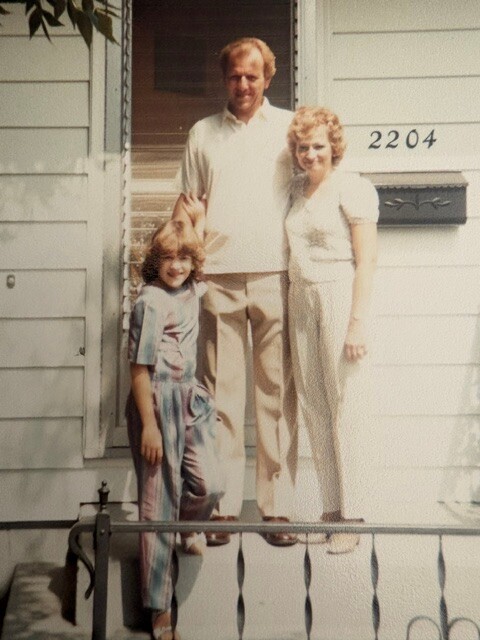 A family of three stands on the front steps of a white house; a man, a woman, and a young girl, with house number 2204 visible.