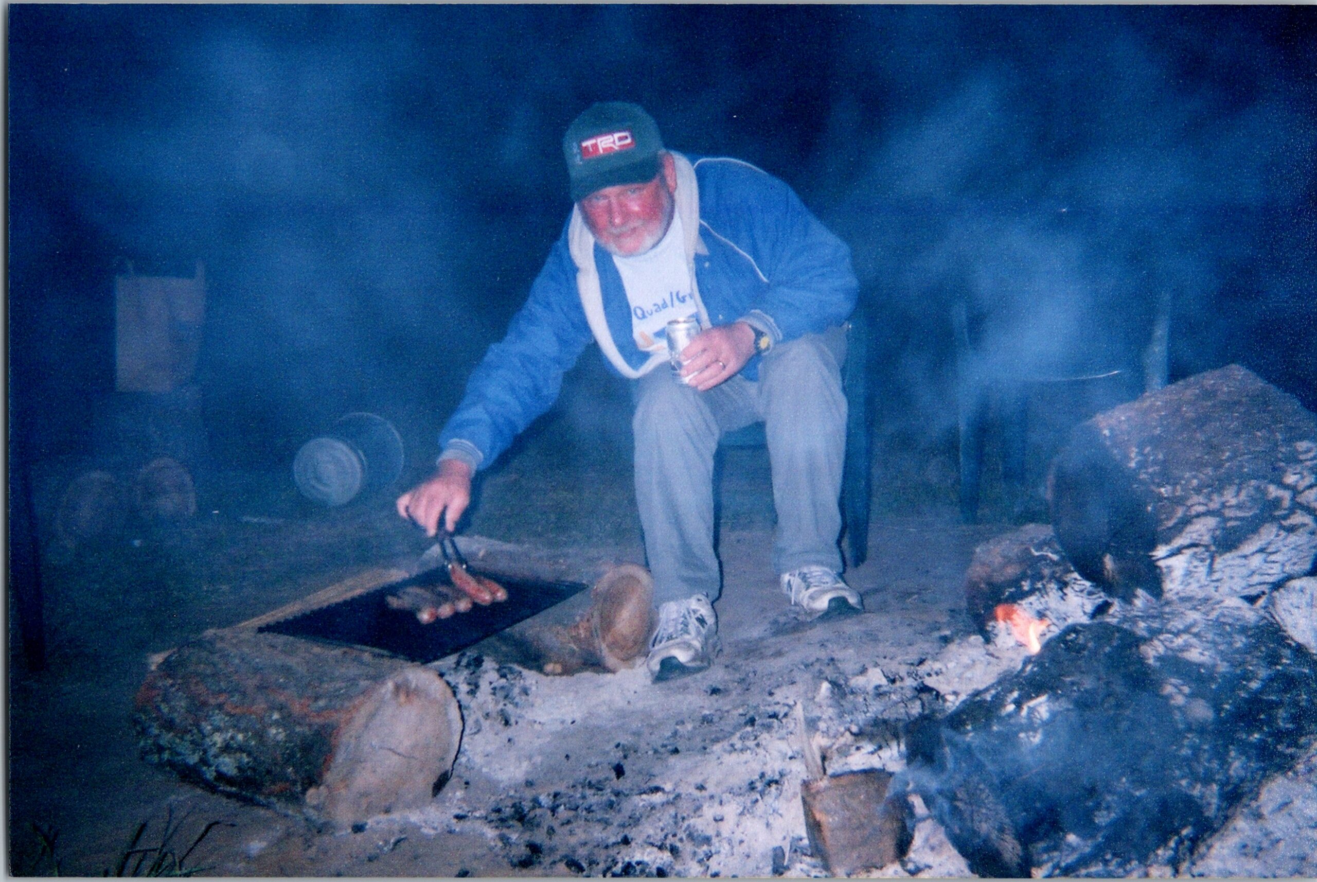 Older man in a blue jacket and TRD cap crouches beside a makeshift outdoor grill, cooking over a smoky fire.
